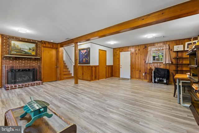 living area with wood walls, stairs, a brick fireplace, beam ceiling, and light wood-style floors