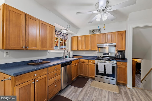 kitchen with dark countertops, under cabinet range hood, a sink, light wood finished floors, and appliances with stainless steel finishes