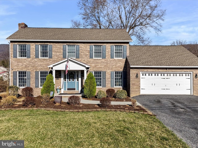 colonial-style house with driveway, an attached garage, a chimney, a front lawn, and brick siding