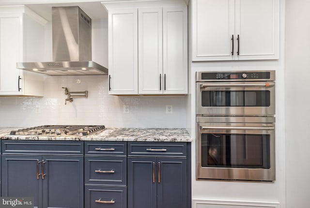 kitchen with white cabinetry, decorative backsplash, blue cabinetry, wall chimney range hood, and appliances with stainless steel finishes