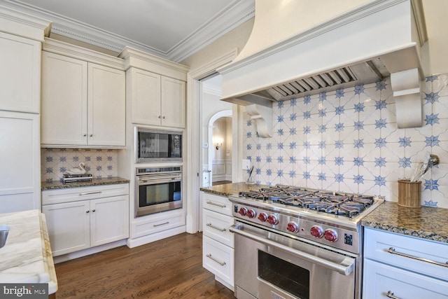 kitchen with crown molding, custom range hood, dark stone counters, dark wood-style floors, and appliances with stainless steel finishes