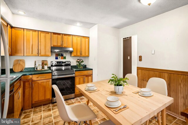 kitchen featuring stainless steel range with electric cooktop, under cabinet range hood, dark countertops, a wainscoted wall, and brown cabinetry