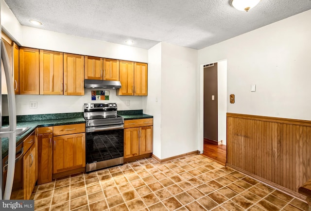kitchen with stainless steel range with electric cooktop, under cabinet range hood, a textured ceiling, dark countertops, and brown cabinetry