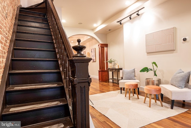 stairs featuring baseboards, arched walkways, brick wall, wood finished floors, and rail lighting
