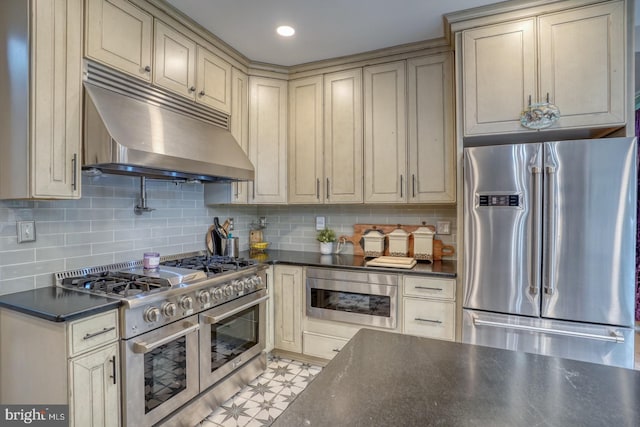 kitchen with appliances with stainless steel finishes, under cabinet range hood, cream cabinets, dark countertops, and tasteful backsplash