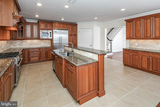 kitchen featuring light tile patterned floors, a sink, an island with sink, built in appliances, and brown cabinetry