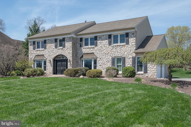colonial home featuring roof with shingles, a front lawn, and stone siding