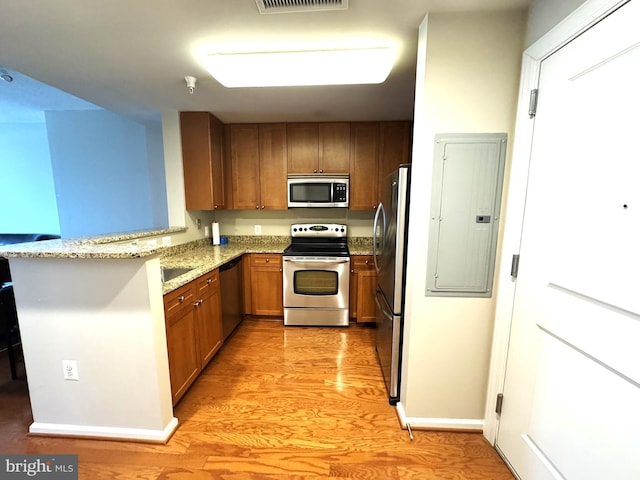 kitchen with brown cabinetry, light stone counters, electric panel, light wood-style floors, and appliances with stainless steel finishes