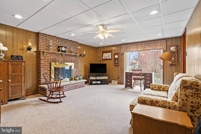 carpeted living room with a brick fireplace, wood walls, a ceiling fan, a paneled ceiling, and a wall mounted AC