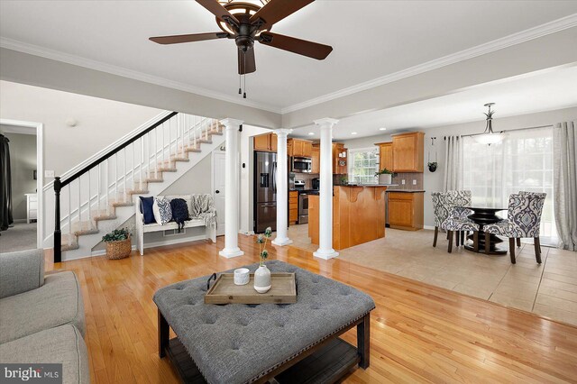living room featuring crown molding, a ceiling fan, stairs, light wood-style floors, and decorative columns