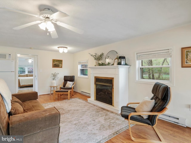 living room featuring baseboard heating, a ceiling fan, a glass covered fireplace, and light wood-style flooring
