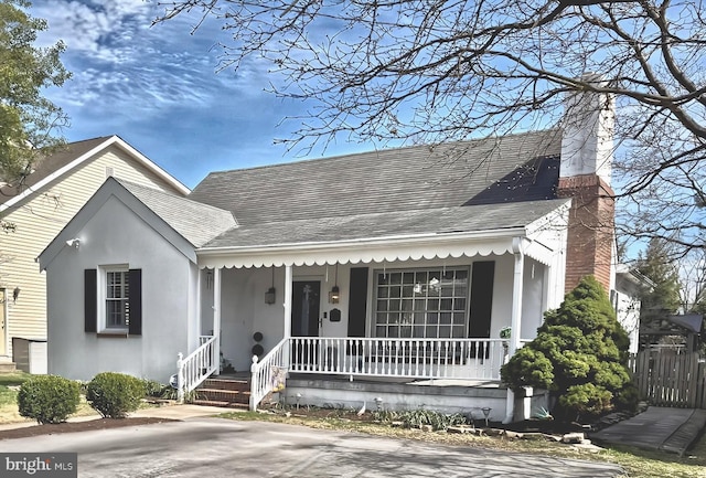 view of front facade featuring roof with shingles, covered porch, a chimney, and stucco siding