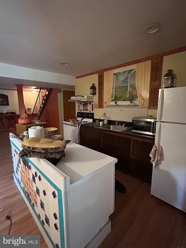 kitchen with white appliances, a sink, wood finished floors, under cabinet range hood, and dark countertops