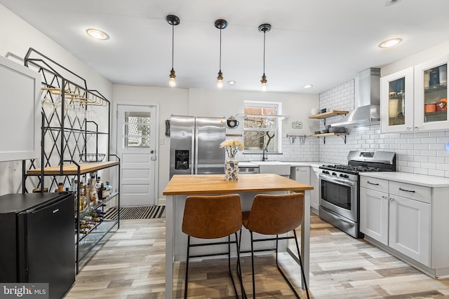 kitchen with backsplash, wall chimney exhaust hood, light wood-style flooring, and appliances with stainless steel finishes