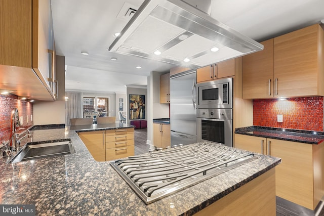 kitchen featuring a peninsula, a sink, exhaust hood, built in appliances, and backsplash