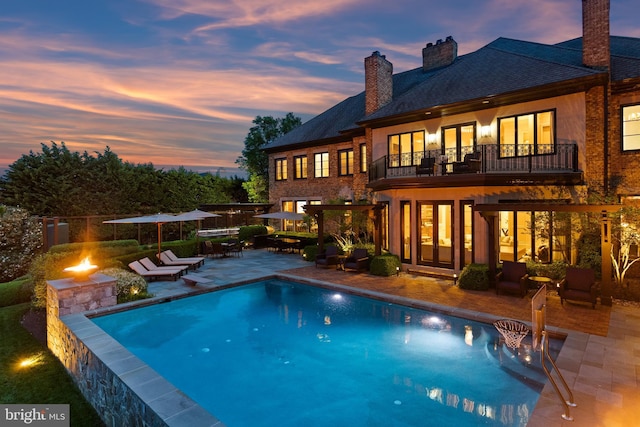 back of property at dusk featuring a fire pit, a balcony, french doors, a chimney, and a patio area