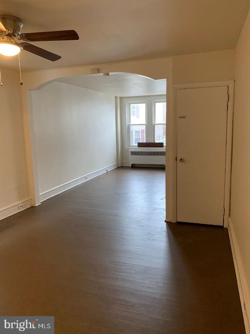 spare room with radiator, baseboards, ceiling fan, arched walkways, and dark wood finished floors