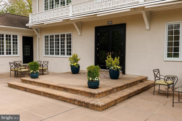 view of exterior entry featuring a patio area, roof with shingles, a balcony, and stucco siding