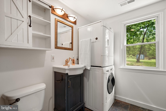 bathroom with stacked washer / drying machine, vanity, toilet, stone finish flooring, and baseboards
