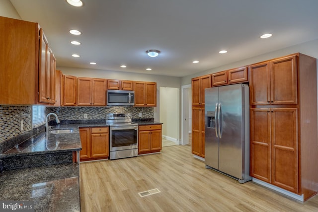 kitchen with appliances with stainless steel finishes, light wood-style floors, a sink, decorative backsplash, and dark stone counters