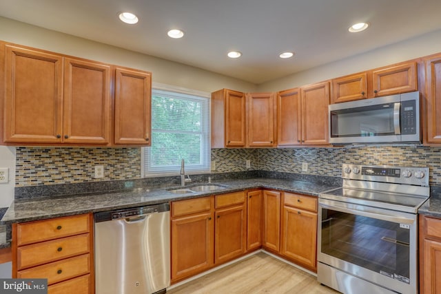 kitchen with stainless steel appliances, a sink, light wood finished floors, dark stone counters, and tasteful backsplash