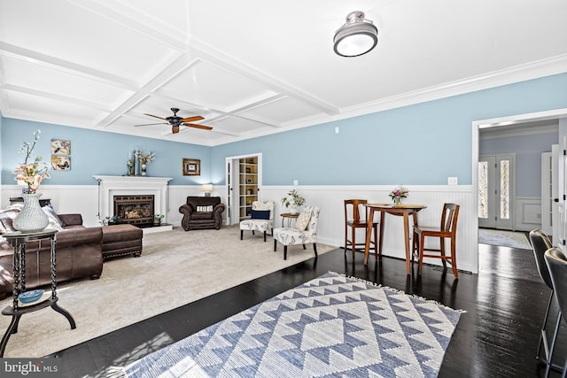 living room with dark wood finished floors, a ceiling fan, a glass covered fireplace, a wainscoted wall, and coffered ceiling