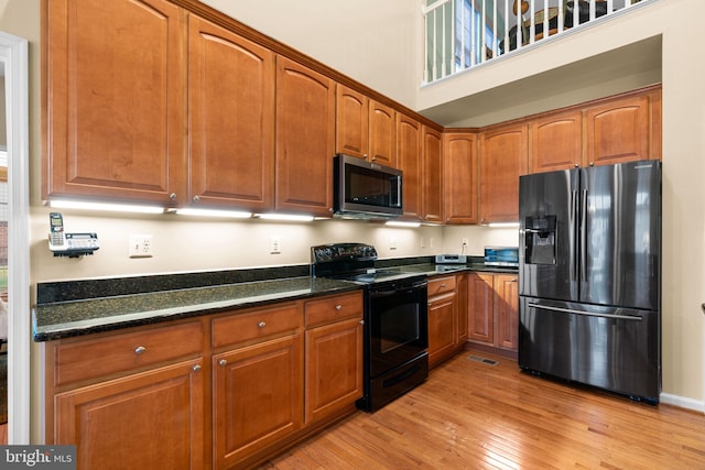 kitchen featuring dark stone counters, light wood-style flooring, a towering ceiling, black appliances, and brown cabinets