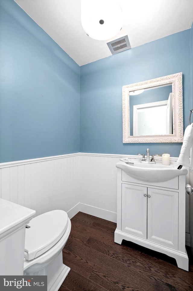 half bathroom with wood finished floors, vanity, a wainscoted wall, toilet, and visible vents