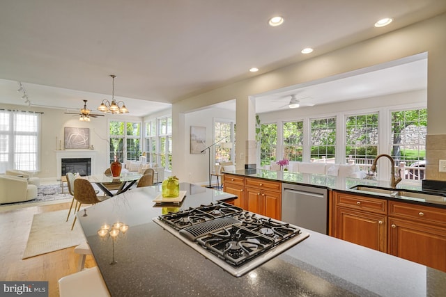 kitchen with stovetop with downdraft, stainless steel dishwasher, brown cabinets, a sink, and ceiling fan with notable chandelier