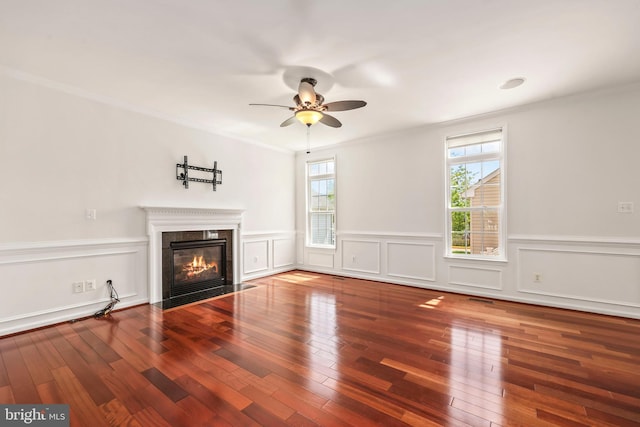 unfurnished living room featuring ceiling fan, a fireplace, wood finished floors, a wainscoted wall, and ornamental molding