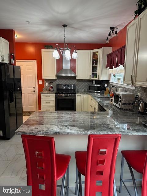kitchen with black fridge with ice dispenser, wall chimney range hood, a peninsula, gas stove, and decorative backsplash