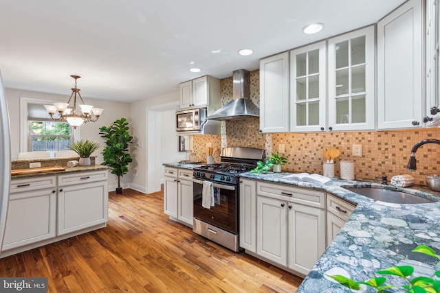 kitchen with a sink, wall chimney range hood, appliances with stainless steel finishes, glass insert cabinets, and light wood-style floors