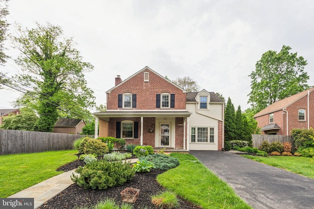 traditional home featuring covered porch, a chimney, fence, a front lawn, and brick siding