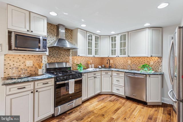 kitchen featuring stone countertops, stainless steel appliances, wall chimney exhaust hood, light wood finished floors, and decorative backsplash