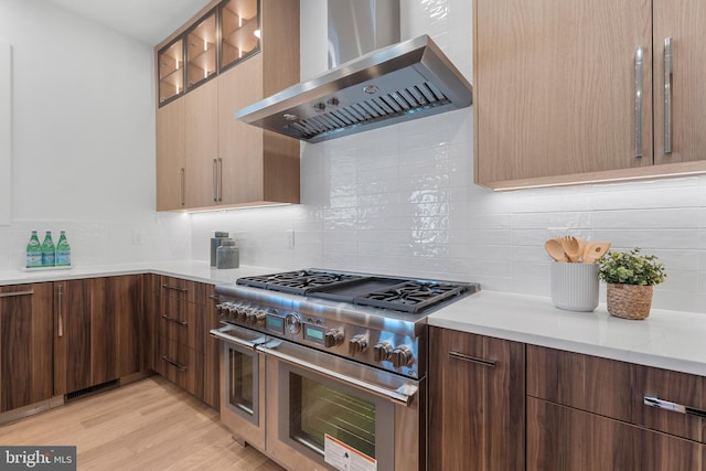 kitchen with double oven range, decorative backsplash, light wood finished floors, wall chimney range hood, and modern cabinets