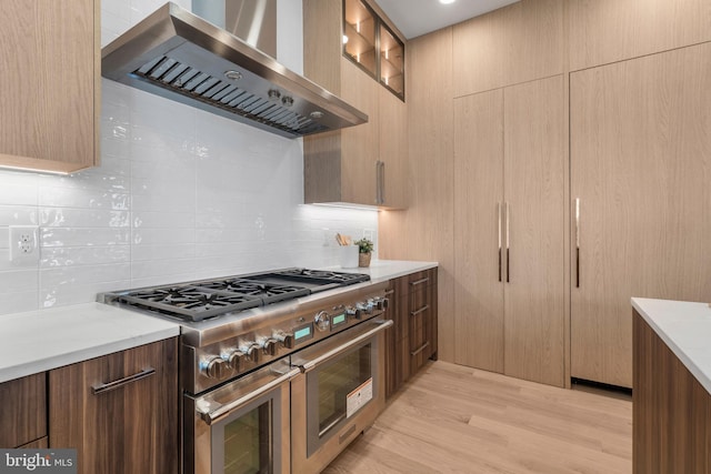 kitchen featuring range with two ovens, wall chimney exhaust hood, light wood-style floors, and decorative backsplash