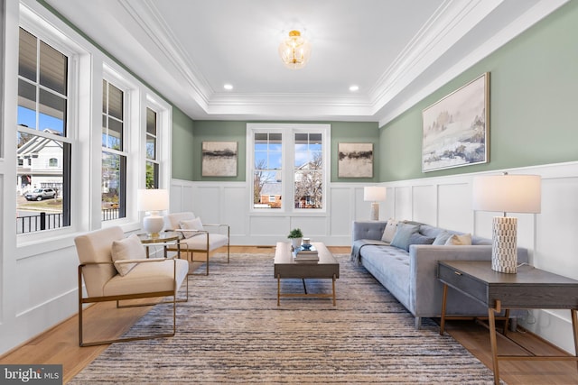 living room featuring wood finished floors, a tray ceiling, ornamental molding, a decorative wall, and wainscoting