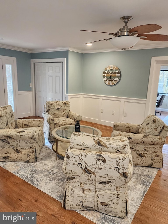 living area with crown molding, a decorative wall, ceiling fan, and wainscoting