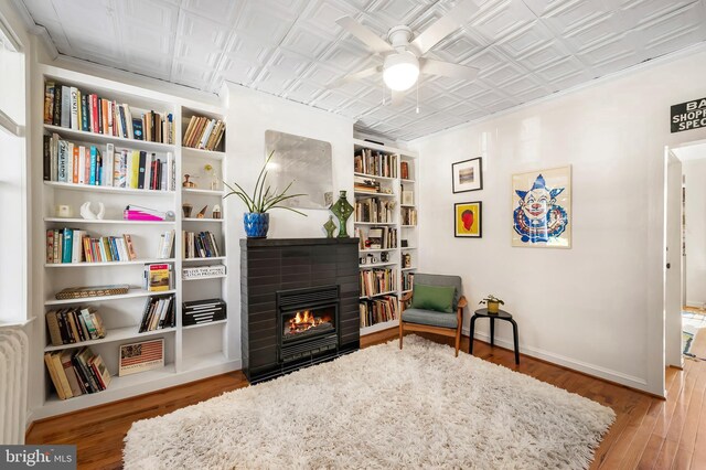 living area with a ceiling fan, wood finished floors, an ornate ceiling, a glass covered fireplace, and baseboards