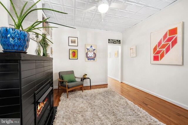 living area featuring wood finished floors, baseboards, an ornate ceiling, ceiling fan, and a lit fireplace