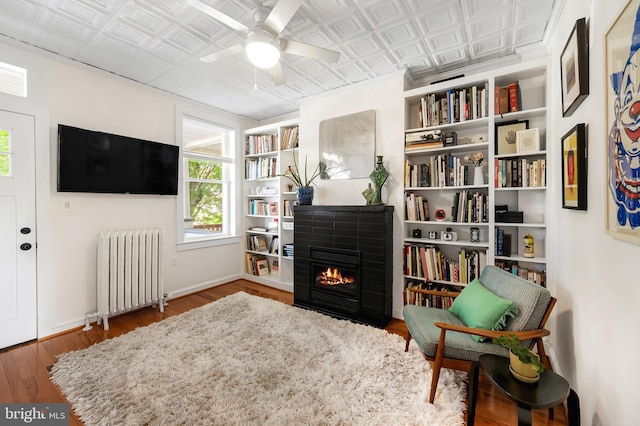 sitting room with radiator, an ornate ceiling, wood finished floors, a lit fireplace, and a ceiling fan