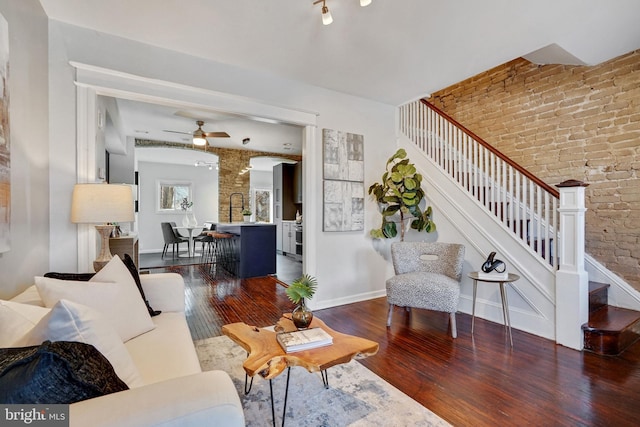 living area featuring a ceiling fan, brick wall, hardwood / wood-style flooring, stairway, and baseboards