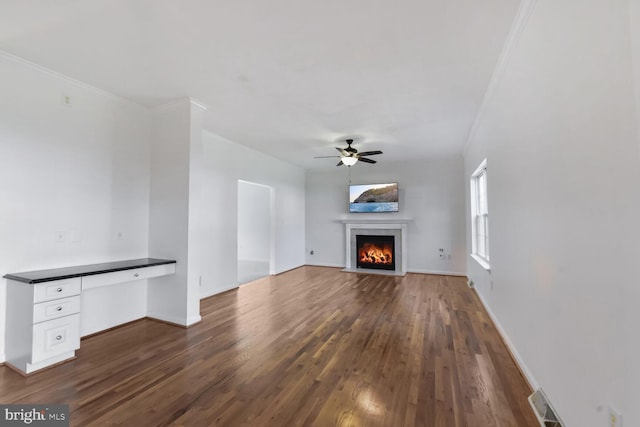 unfurnished living room with built in desk, a ceiling fan, crown molding, a fireplace with flush hearth, and dark wood-type flooring