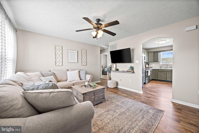 living room with a textured ceiling, dark wood-style floors, baseboards, arched walkways, and ceiling fan