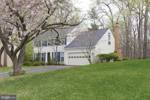 colonial-style house featuring aphalt driveway, roof with shingles, an attached garage, a front lawn, and a chimney