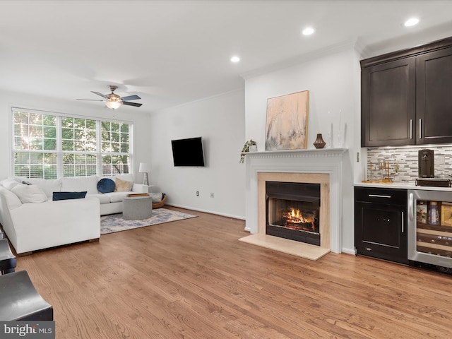 living room featuring indoor wet bar, beverage cooler, a lit fireplace, a ceiling fan, and light wood-style flooring