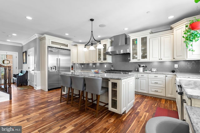 kitchen with crown molding, dark wood finished floors, a center island with sink, wall chimney range hood, and appliances with stainless steel finishes