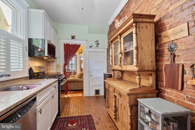 kitchen with appliances with stainless steel finishes, a sink, brick floor, white cabinetry, and a wealth of natural light