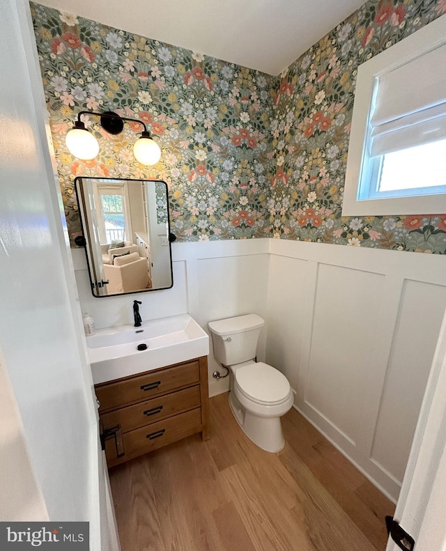 bathroom with wallpapered walls, wood finished floors, wainscoting, vanity, and toilet