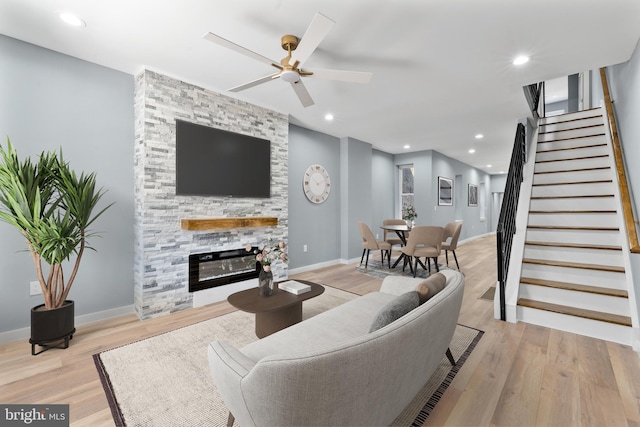 living area with stairs, light wood-style flooring, a ceiling fan, and a stone fireplace
